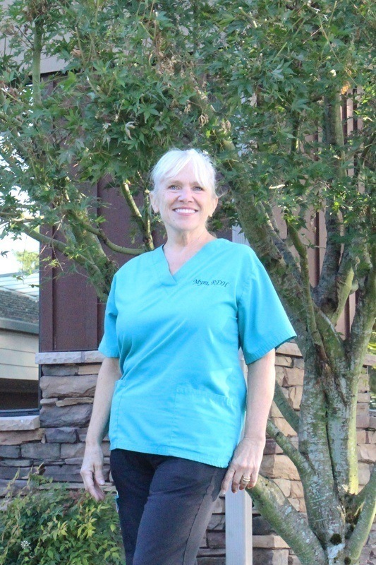 Myra wearing an aqua scrub shirt against a nature and building background.